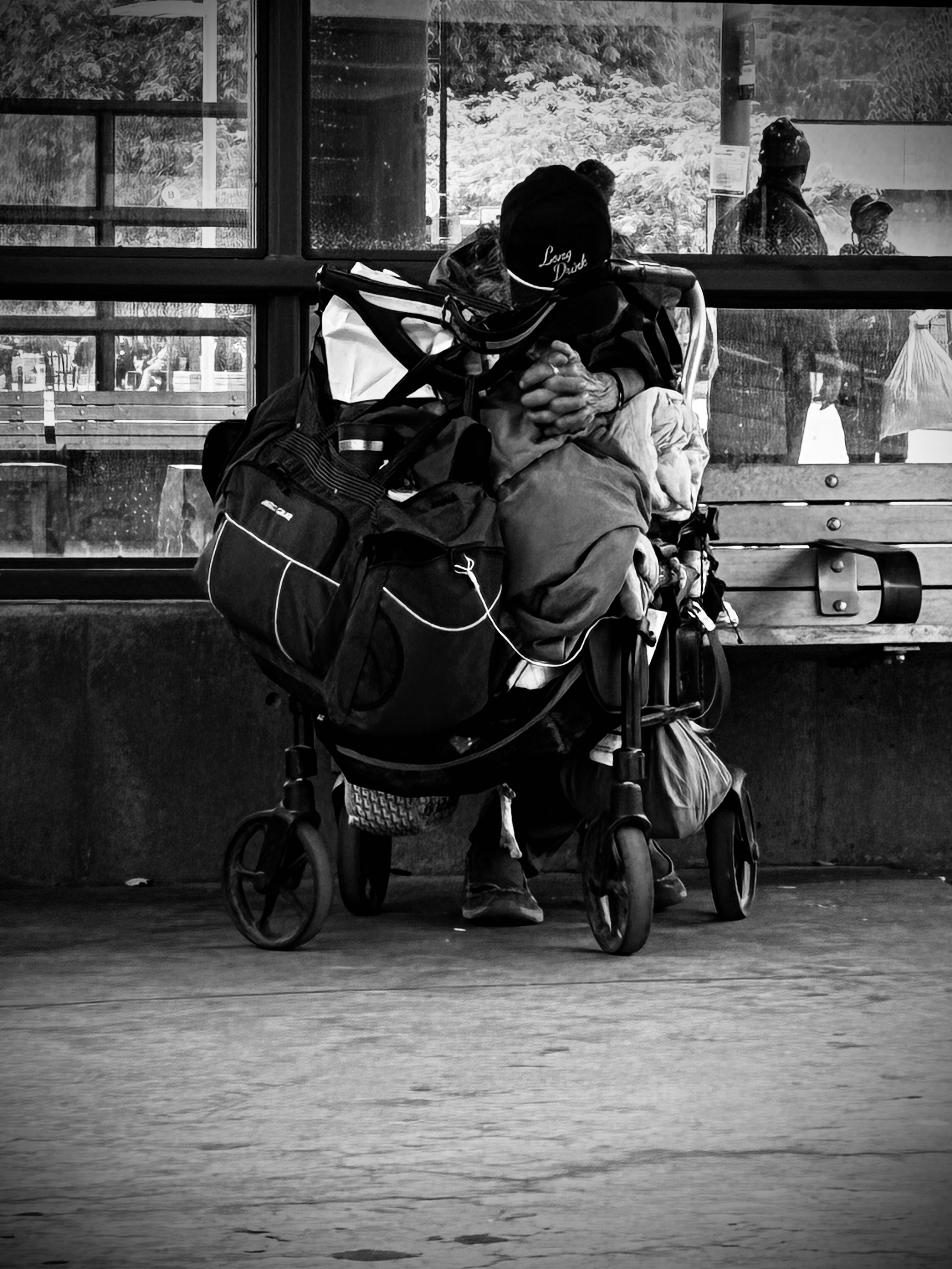 The Third Man, a black-and-white documentary photograph by Randy Millis showing a person seated with a heavily loaded mobility walker at a Kelowna transit stop, hands clasped beneath a baseball cap.
